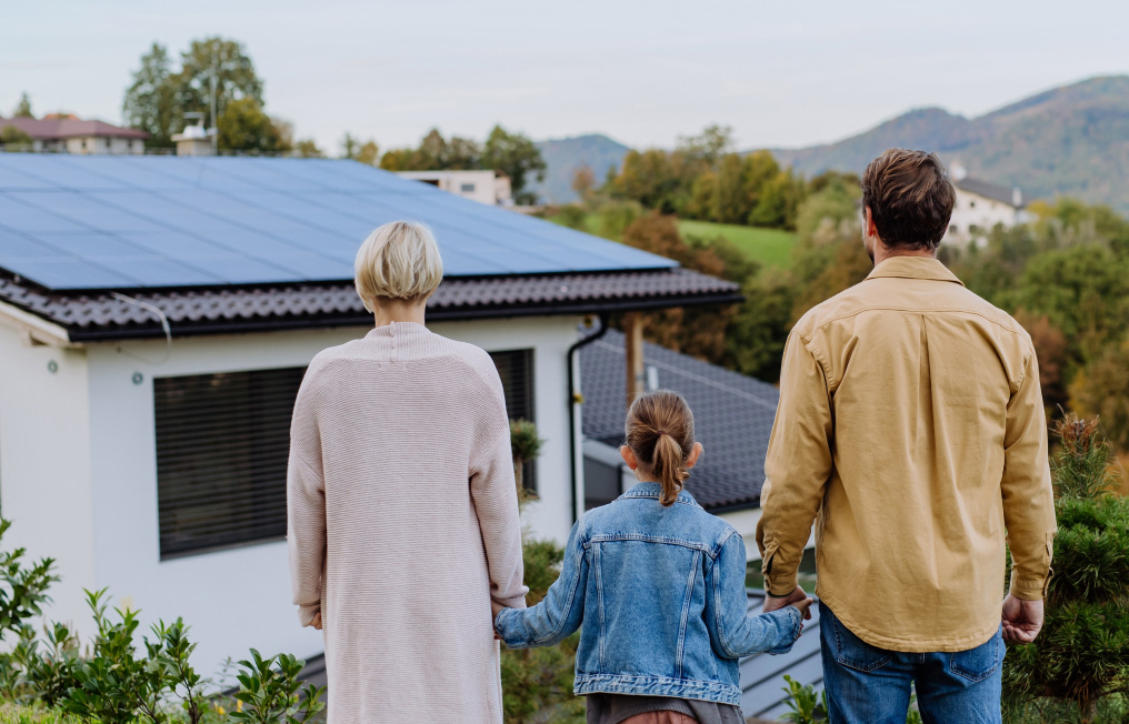 Familie steht vor einem Haus mit Solarpanelen auf dem Dach und genießt die sonnige Umgebung.